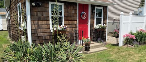 A single-story house with a red front door, two windows, and a porch with a black railing. There are flower pots on the porch and a wreath on the door.