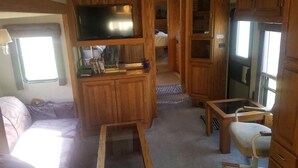 A compact RV interior with a kitchenette, a TV mounted on a wooden cabinet, a small table, and a cushioned seating area. - Montana Mountain Memories- RV in Orchard by Trout Pond (Florence)