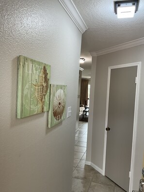 A hallway with two wall-mounted artworks, a door, and a tiled floor leading to a dining area. - Beautiful Beach & Pool Front Condo- Remodeled in March 2024 (Galveston)