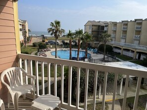 A balcony with white chairs, a swimming pool, palm trees, and a view of the ocean and buildings. - Beautiful Beach & Pool Front Condo- Remodeled in March 2024 (Galveston)