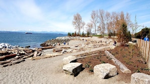 A sandy beach with scattered rocks and driftwood, a wooden fence, and leafless trees. - Waterfront 2BD Next to Pike Place w/ Private Patio (Seattle)