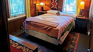 Ein Zimmer mit Holzwänden, einem Bett, zwei Nachttischlampen, einem Teddybären und einem fenster mit Vorhängen.