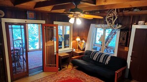 Ein gemütliches Blockhaus-Interieur mit Holzbalkendecke, Deckenventilator, einer schwarzen Couch, einem gemusterten Teppich und einem Essbereich mit Tisch und Stühlen.