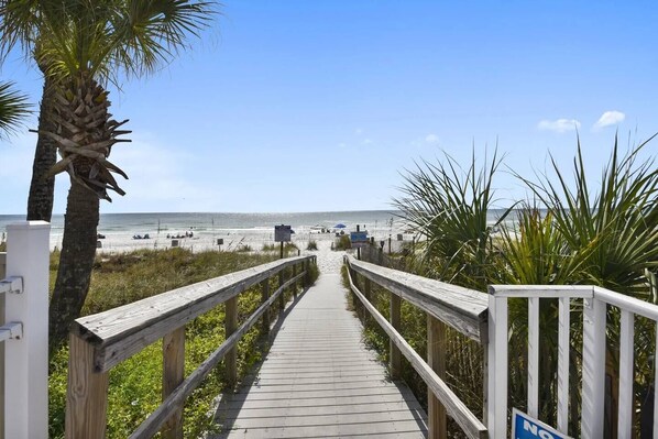 A wooden boardwalk leading to a beach with palm trees and a clear blue sky.