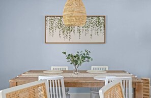 A dining area with a wooden table, white chairs, a woven pendant light, and a framed botanical print on the wall.