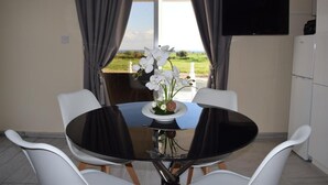 Een eetruimte met een ronde glazen tafel, witte stoelen, een vaas met witte bloemen en een aan de muur bevestigde televisie.