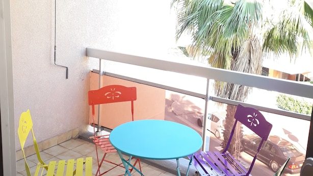 A balcony with a blue table and colorful chairs, a white railing, and a view of palm trees and parked cars. - Studio Avec Clim et Parking Privėe (Cavalaire-sur-Mer)