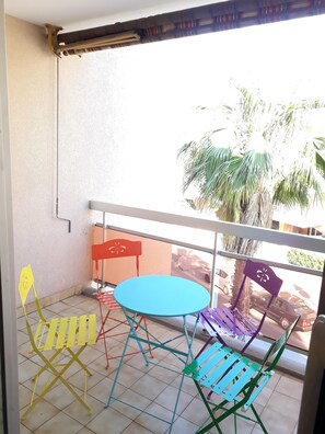 A balcony with a blue table and colorful chairs, a white railing, and a view of palm trees and parked cars.