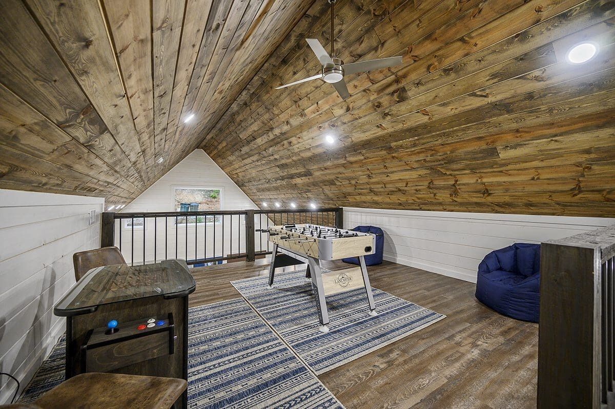 Quarto estilo loft com teto de madeira, mesa de futebol de bolso, puff azul e vista para o exterior.