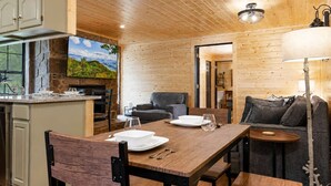 Un interno in legno con un tavolo da pranzo apparecchiato per due persone, un divano e un angolo cottura con parete in pietra e un'ampia finestra.