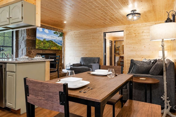 Un interior de madera con una mesa de comedor preparada para dos personas, un sofá y una zona de cocina con una pared de piedra y un ventanal grande.