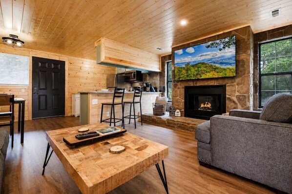 A cozy living room with a fireplace, wooden furniture, and a kitchen area.
