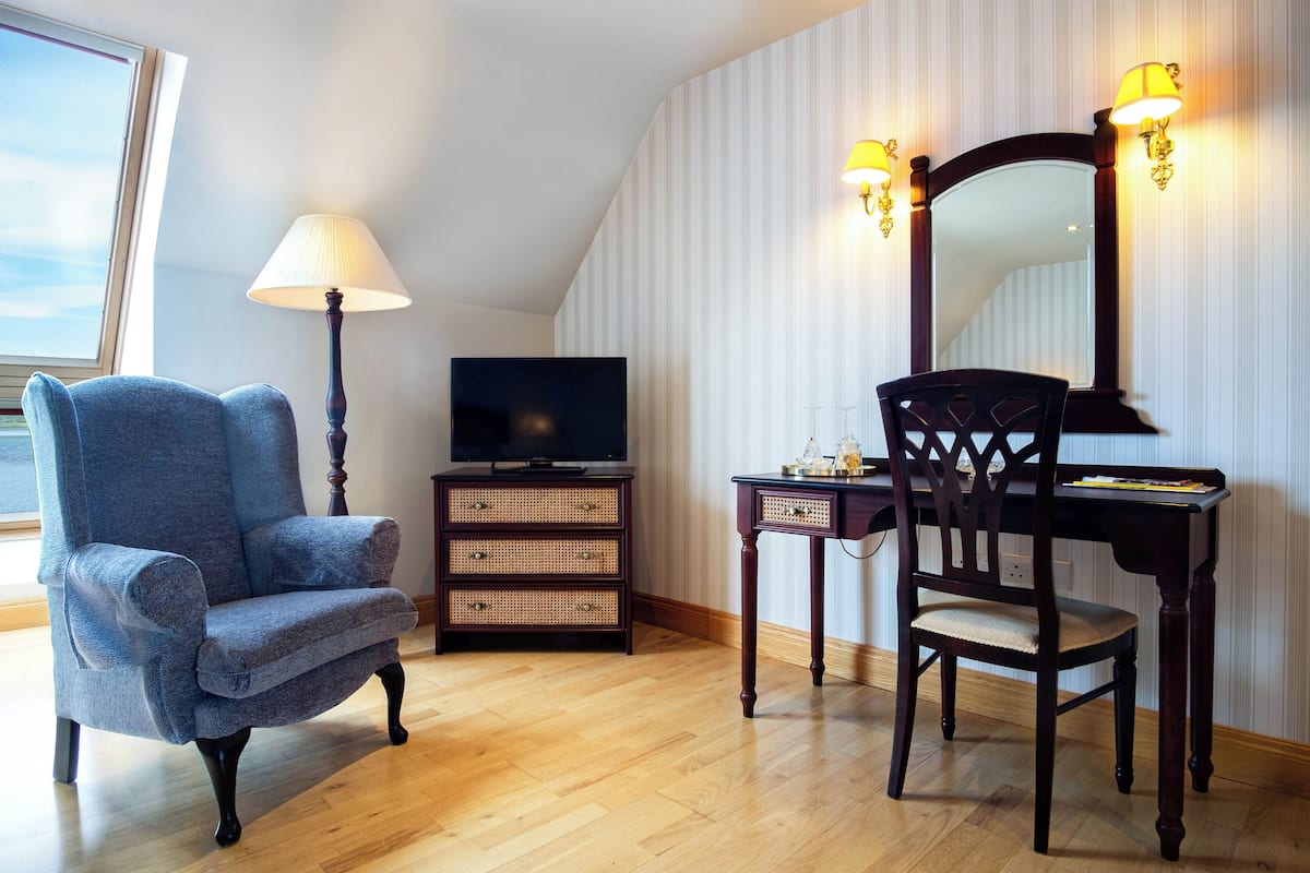 A room with a blue armchair, a wooden desk with a chair, a television, and a lamp.