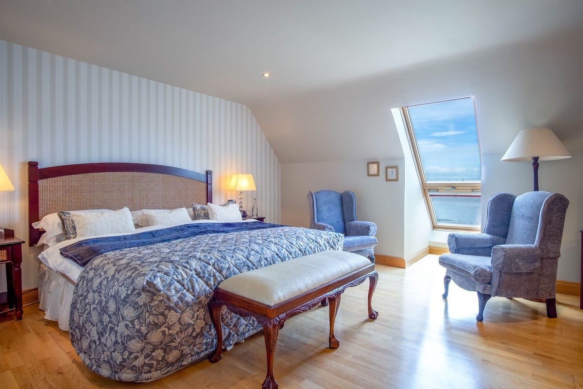 A bedroom with a large bed, two armchairs, a bench, a nightstand, a lamp, and a window with a view of the sea.