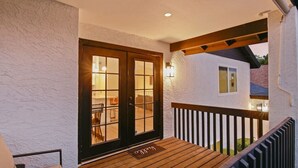 Une terrasse en bois avec une rambarde noire, une porte dotée de panneaux de verre et une petite fenêtre sur le côté d’un bâtiment.