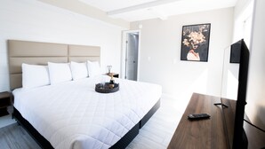 A modern hotel room with a large bed, a bedside table with a remote control, a framed picture on the wall, and a door leading to another room. - 300 Tenth (Myrtle Beach)