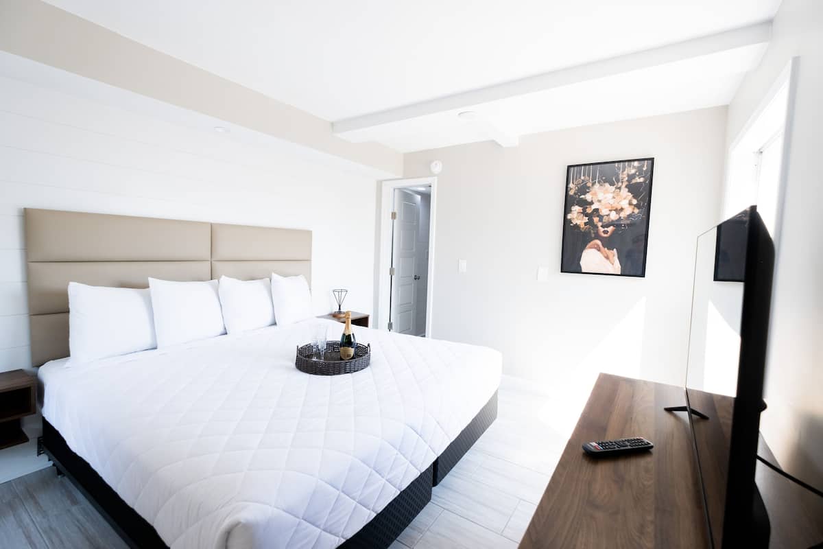A modern hotel room with a large bed, a bedside table with a remote control, a framed picture on the wall, and a door leading to another room.