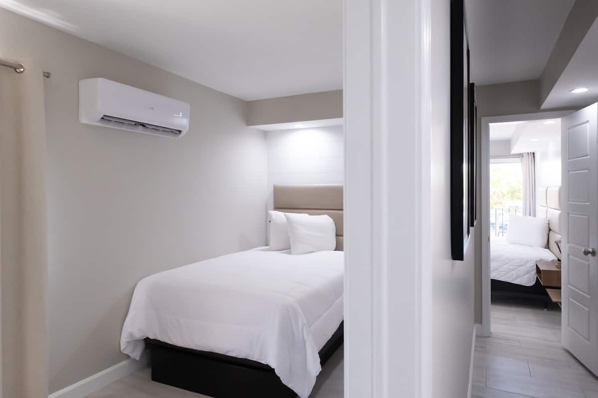 A modern bedroom with a bed, an air conditioning unit, and a view of another room through an open door.