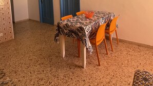 En matplats med ett bord täckt av en bordsduk med ett lövmönster, omgivet av orangefärgade stolar. Det finns två blå dörrar och ett konstverk som hänger på väggen.