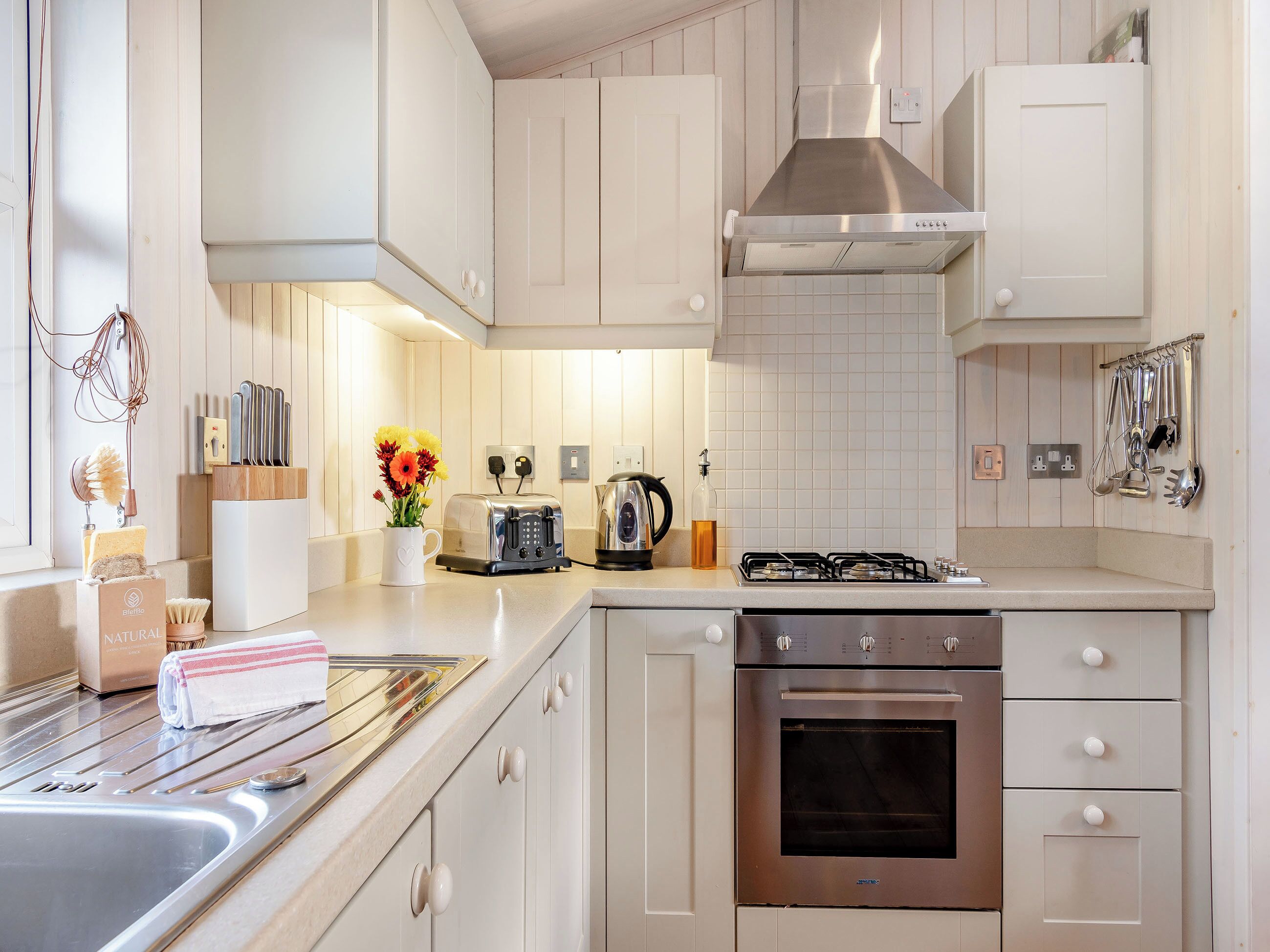 A modern kitchen with white cabinetry, a stainless steel range hood, a gas stove, a built-in oven, and a countertop with a toaster and a kettle.