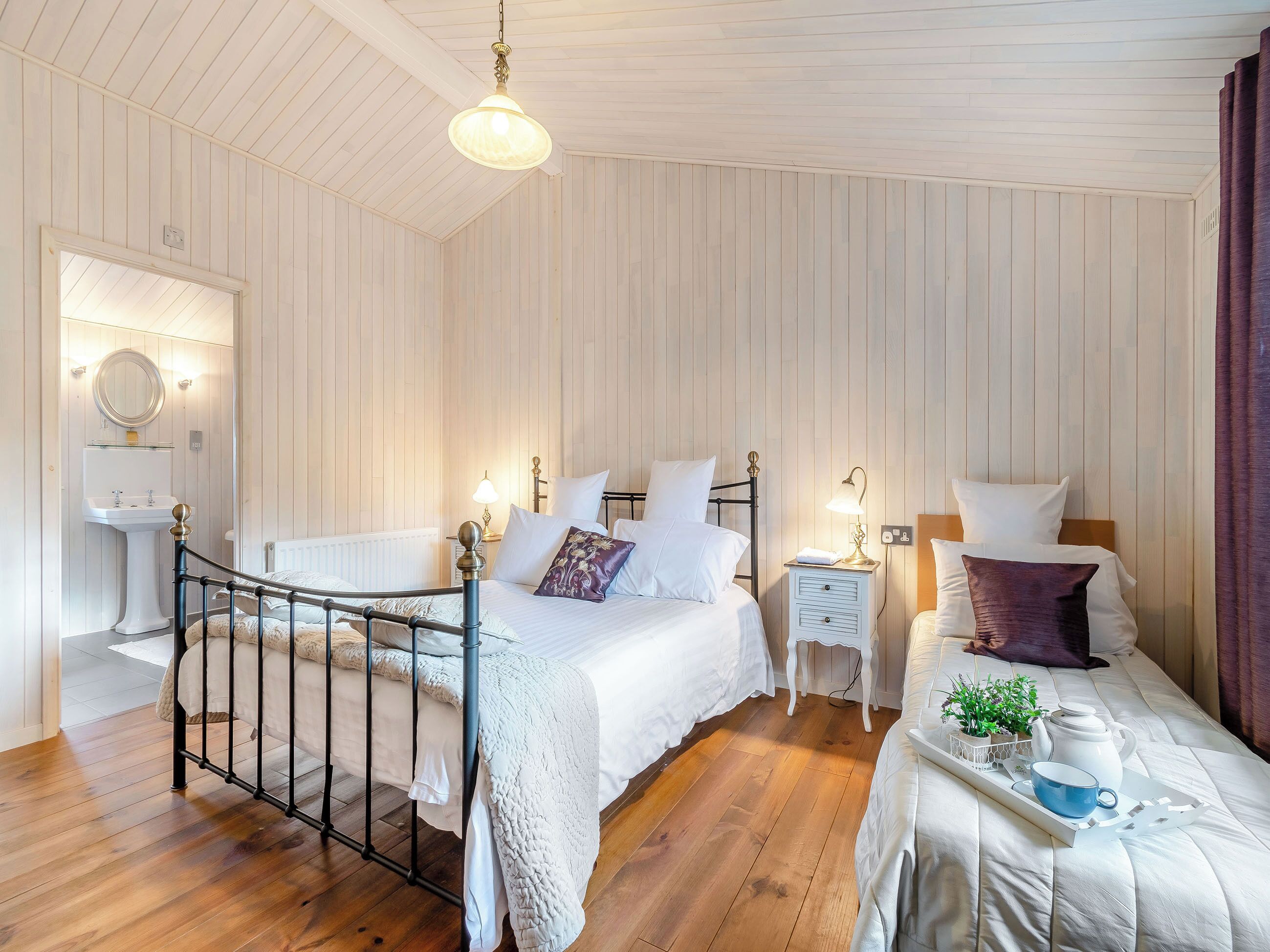 A bedroom with a wooden floor, a bed with white bedding, a bedside table with a lamp, and a small window.