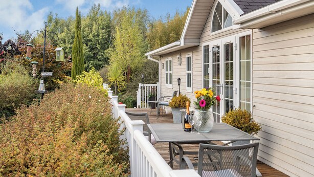 Une maison avec une terrasse, une table à manger et des chaises. Sur la table, on trouve une bouteille de champagne et un vase rempli de fleurs.