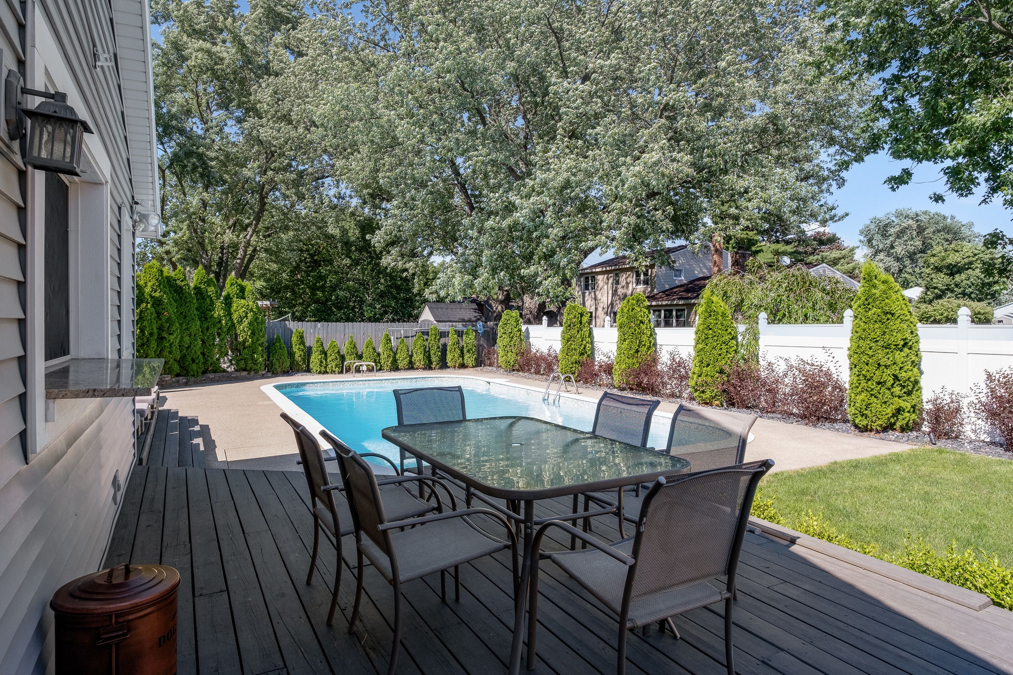 A poolside area with a glass table and chairs, surrounded by a wooden deck and a white fence.