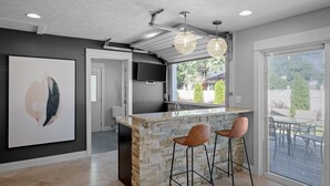 A modern kitchen with a stone island, two bar stools, a flat-screen TV, and a large window overlooking a patio. - Private Pool, Hottub, Steamshower just 10 minutes from Grand Rapids (Grandville)