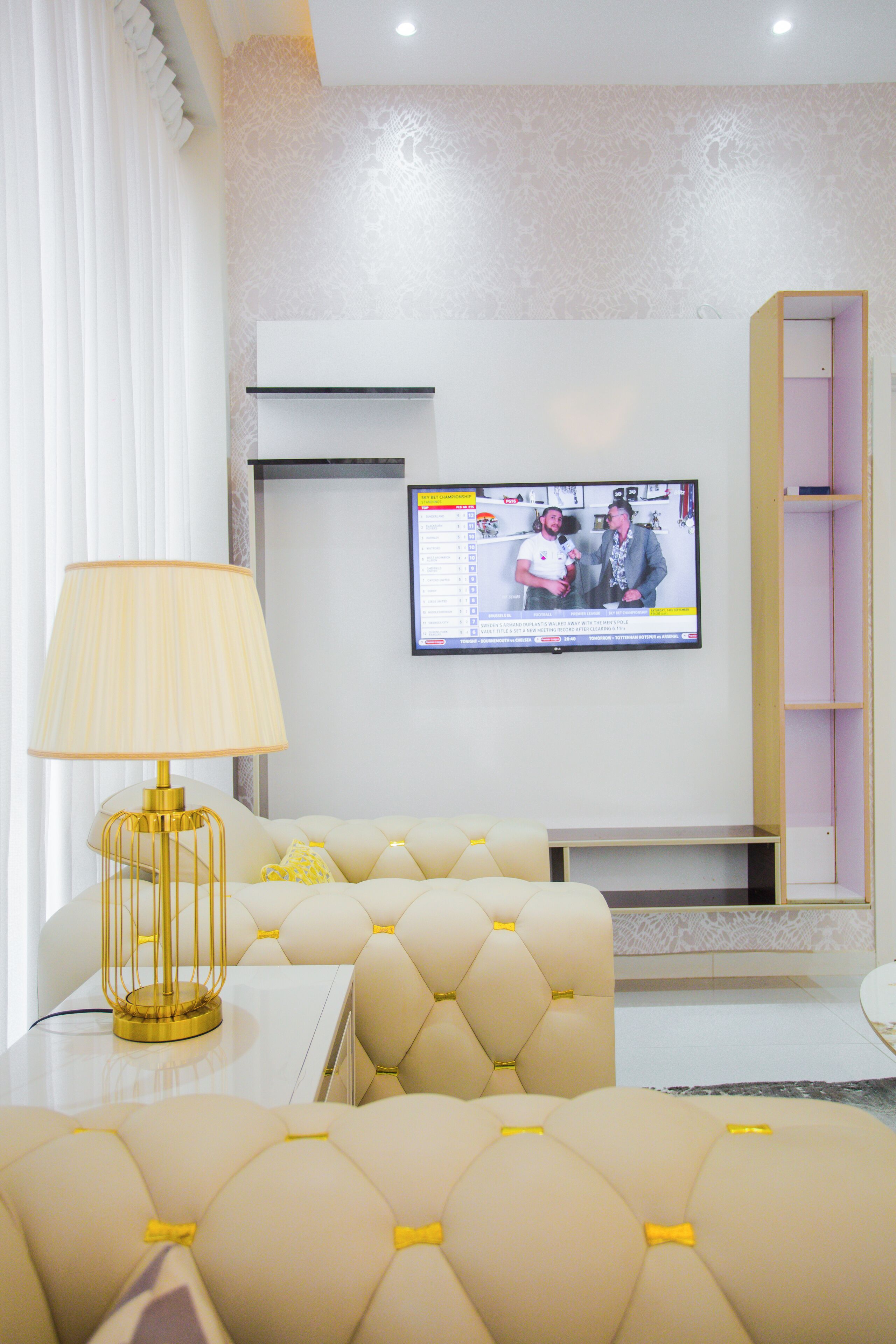 A modern living room with a television mounted on the wall, a sofa with yellow accents, and a gold lamp on a white table.