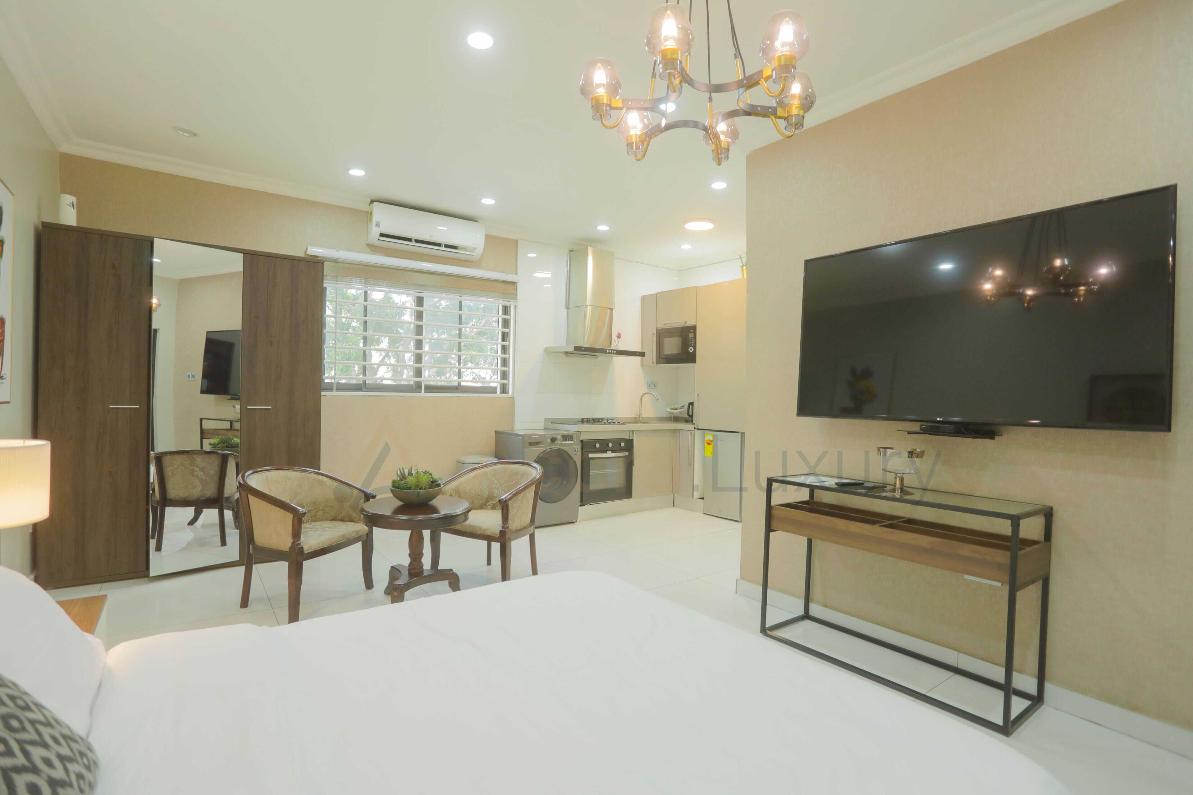 A modern hotel room with a large bed, a dining area with chairs, a small kitchen, and a TV mounted on the wall.