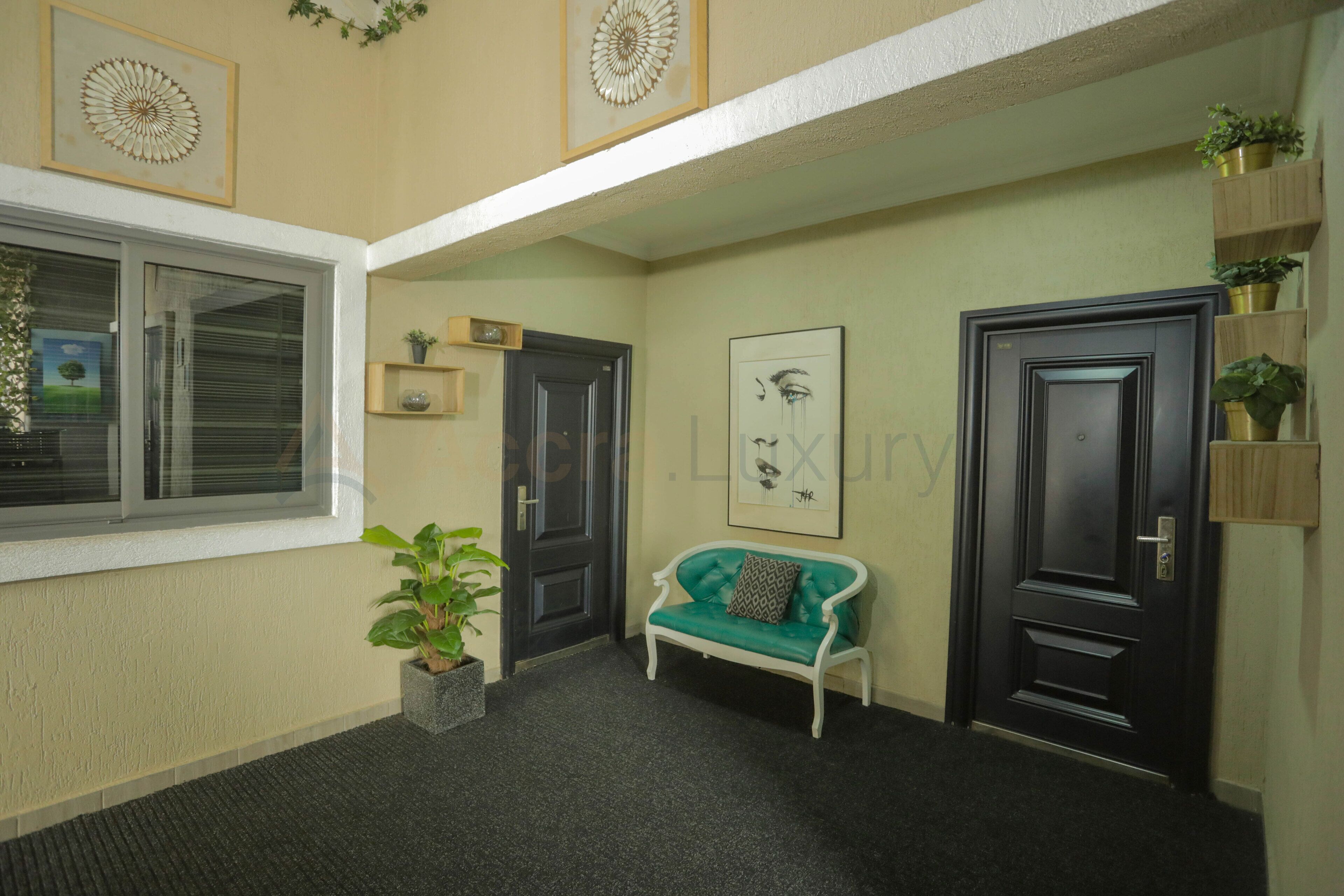 Um corredor com um sofá verde, duas portas pretas, uma janela com persianas e um vaso de planta.