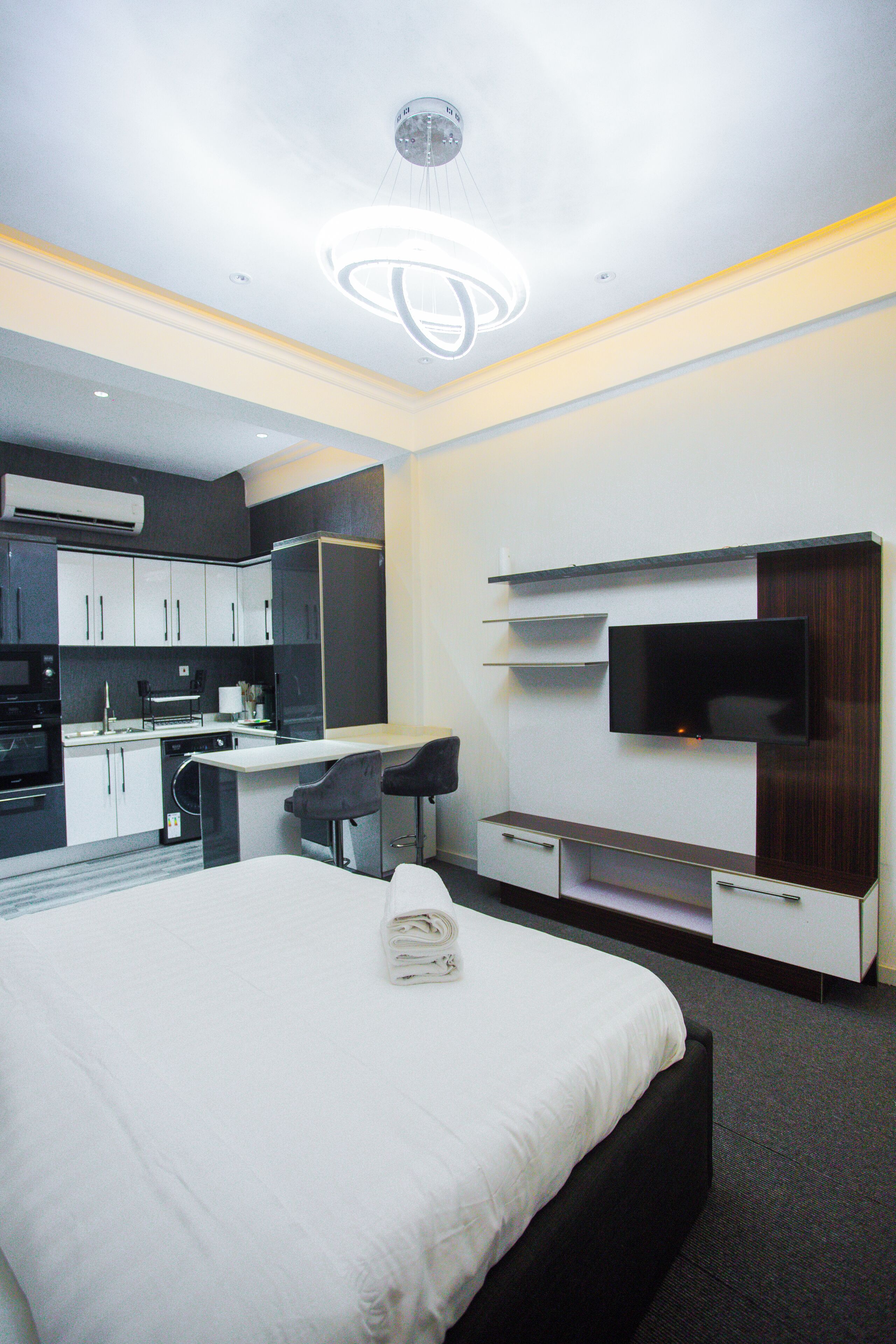 A modern hotel room with a bed, a TV, a kitchen area, and a dining table.