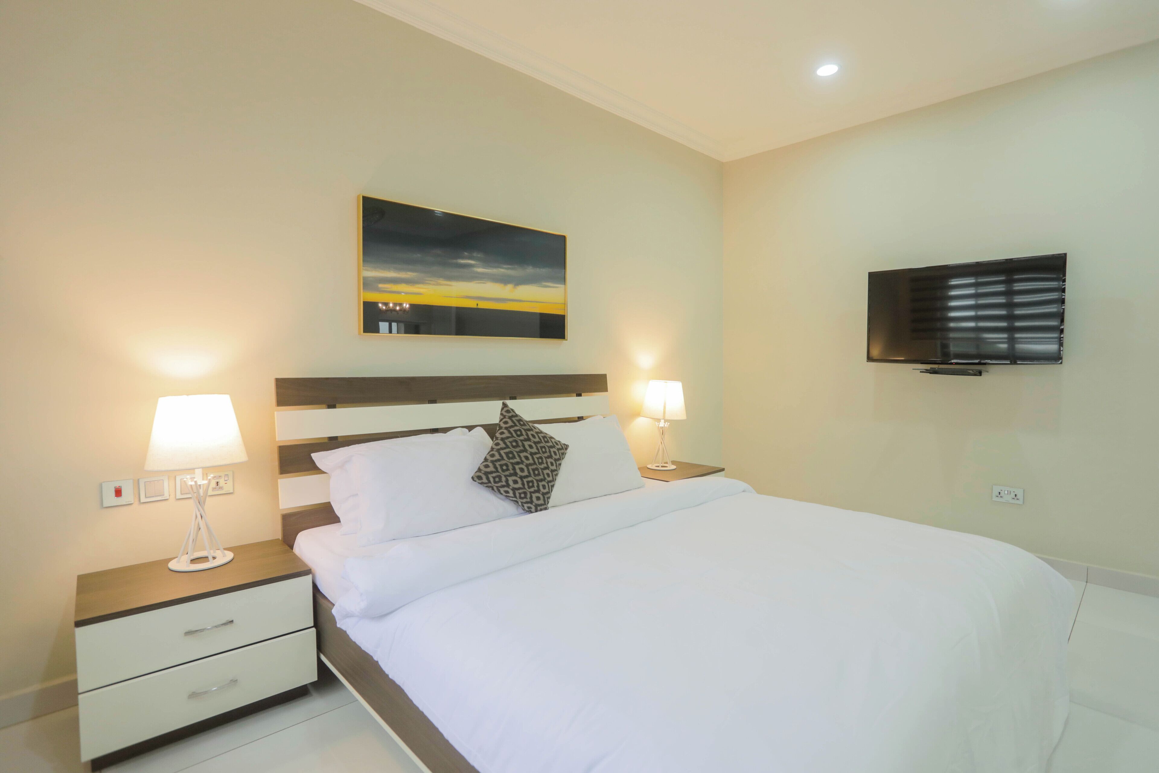 A neatly made bed with white linens, two bedside lamps, a nightstand with drawers, a painting above the bed, and a flat-screen TV on the wall.
