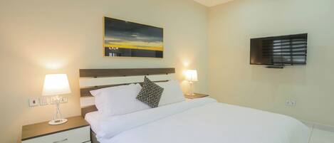 A neatly made bed with white linens, two bedside lamps, a nightstand with drawers, a painting above the bed, and a flat-screen TV on the wall.