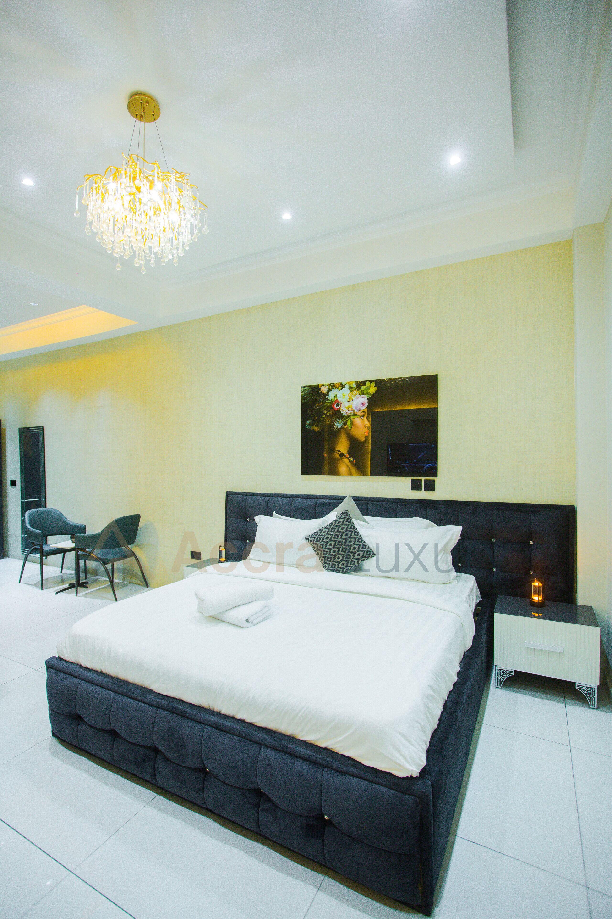 A modern bedroom with a large bed, a chandelier, a chair, and a bedside table.
