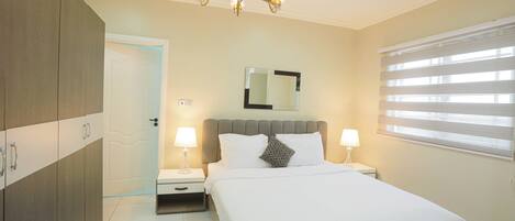 A bedroom with a bed, nightstands, a chandelier, a mirror, and a window with blinds.