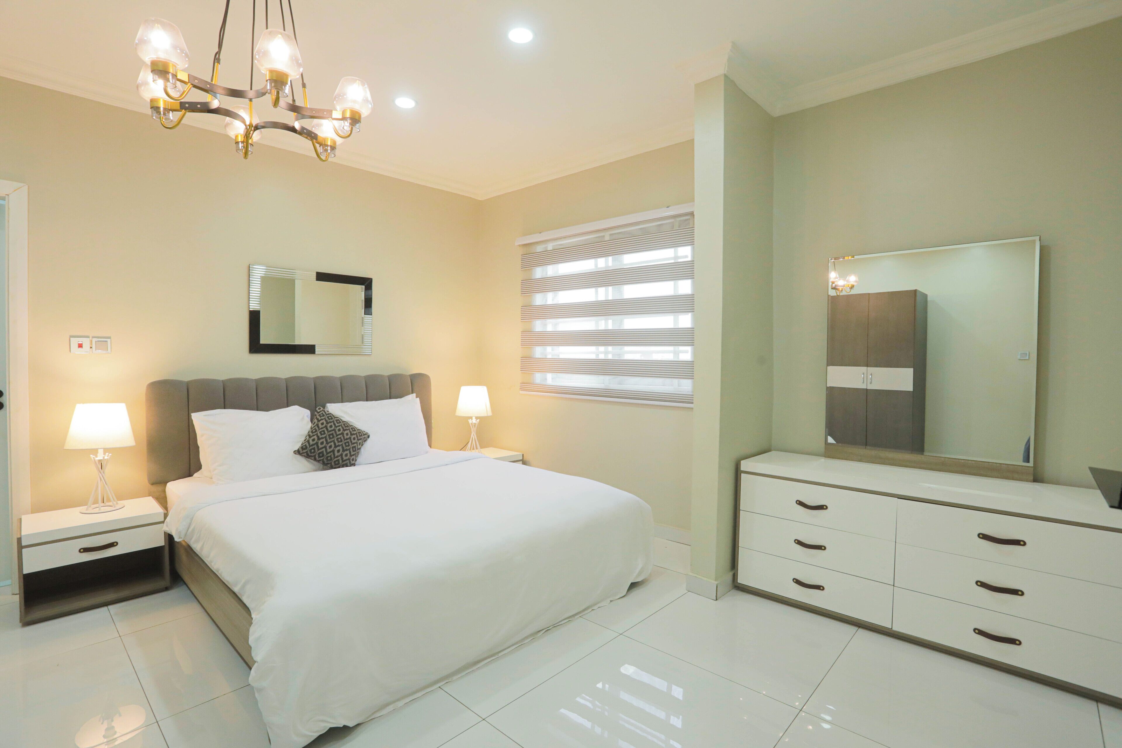 A modern bedroom with a large bed, bedside tables with lamps, a dresser, a mirror, and a chandelier.