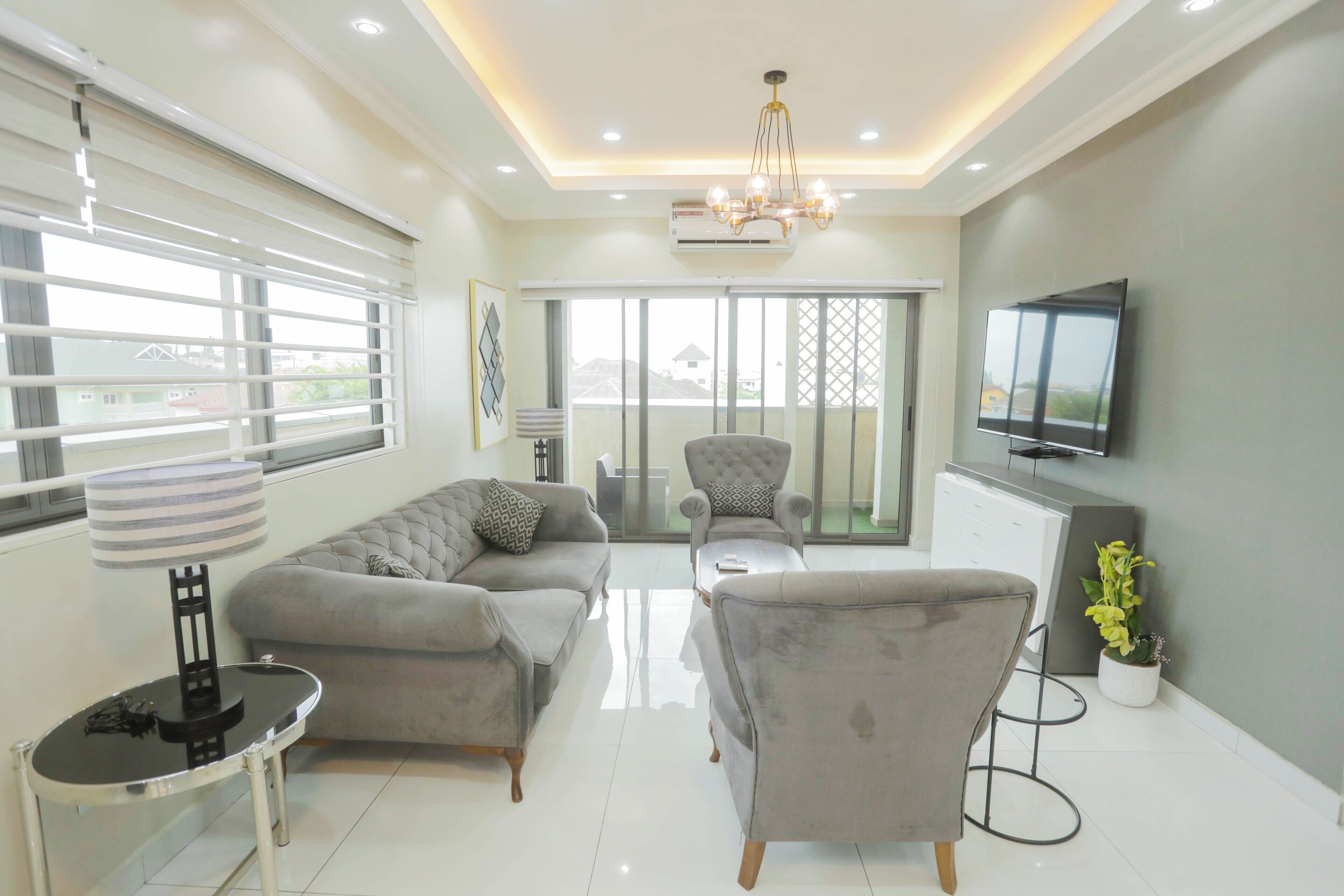 A modern living room with a sofa, armchair, and a small table. There is a TV mounted on the wall and a balcony with a view.