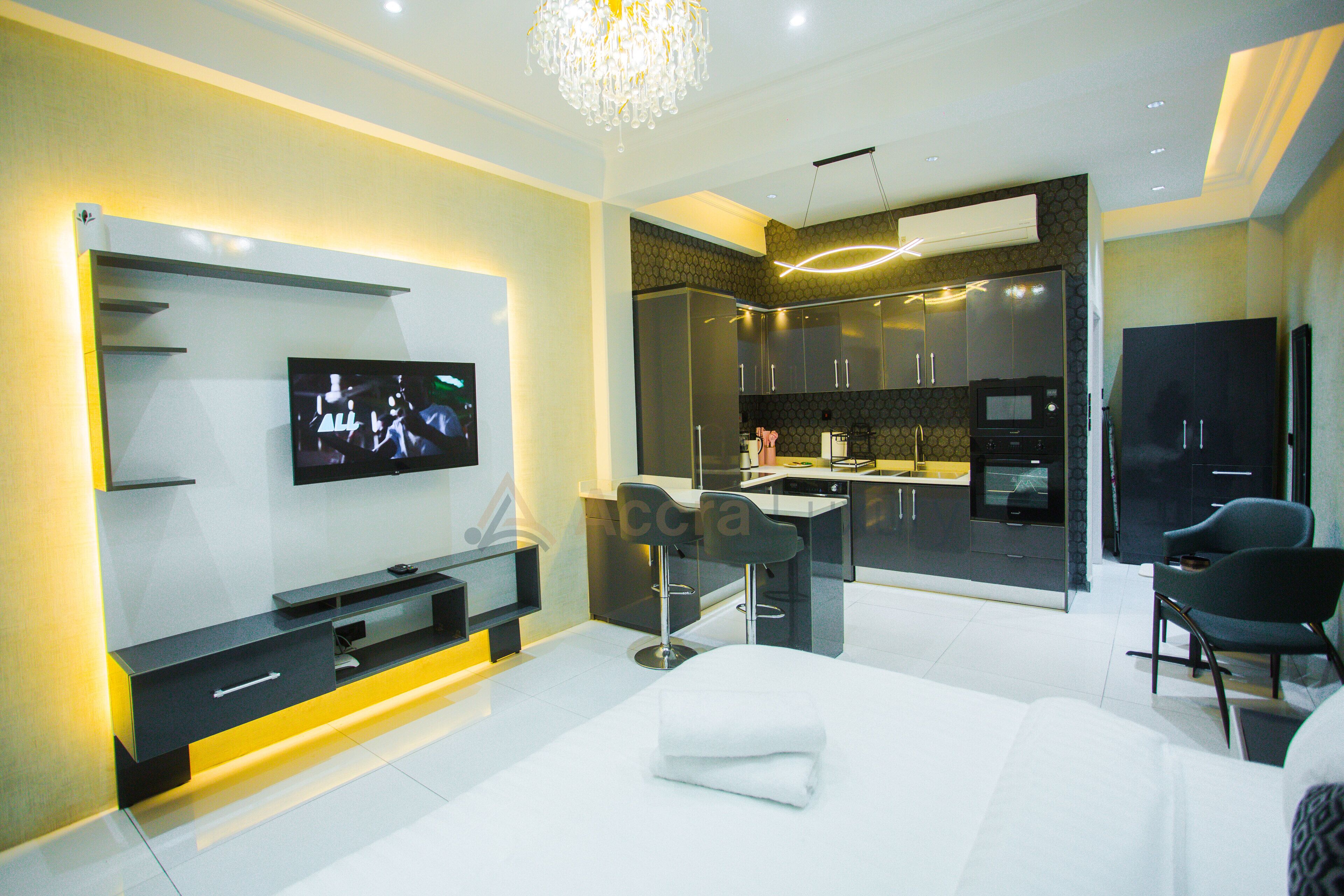 A modern hotel room with a TV, a bed, a seating area, and a kitchenette.