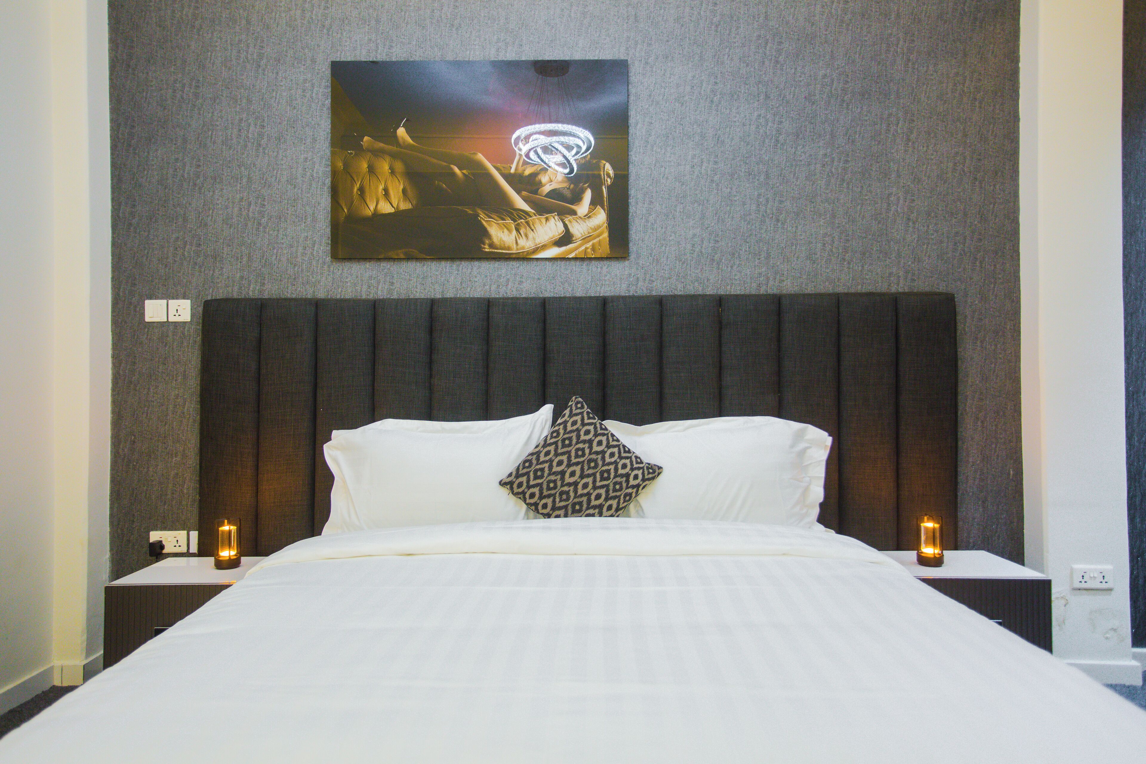 A neatly made bed with white linens, a dark headboard, and a patterned pillow. There is a wall-mounted artwork above the bed and two bedside lamps on either side.