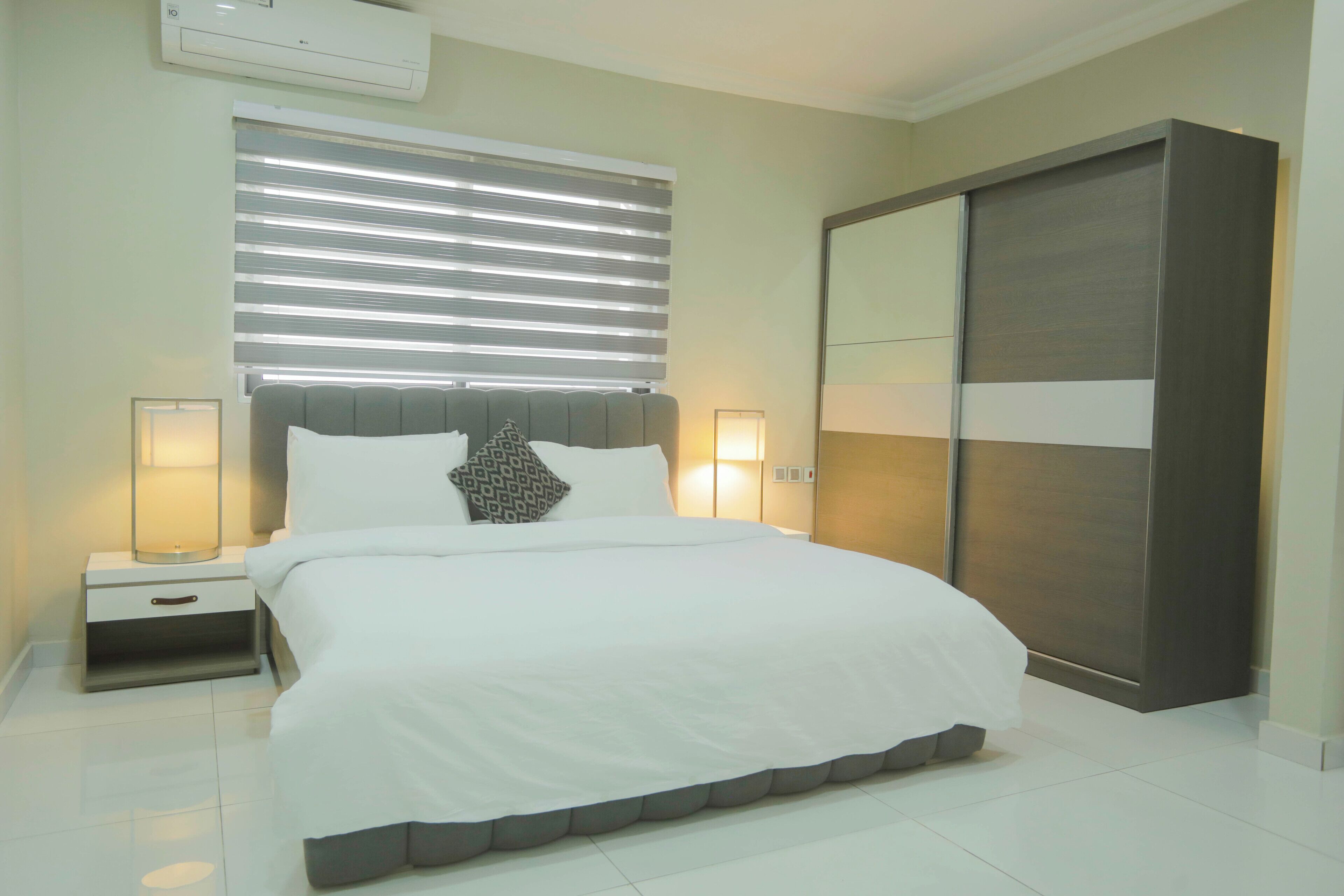 Une chambre à coucher comprenant un lit, des lampes de chevet, une table de chevet, une grande armoire et une fenêtre avec des stores.