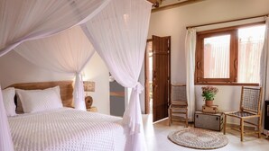 Un dormitorio con cama con dosel, cabecera de madera, mesita de noche con lámpara, ventana con cortinas, alfombra redonda pequeña y una silla.