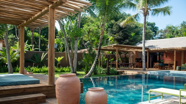 Zona junto a la piscina con escalones de madera, dos macetas grandes de terracota y un área de estar cubierta con mesas y sillas.