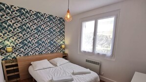 En pænt redt seng med hvide sengetøj og håndklæder, en væg med geometrisk mønster, en sengetavlelampe og et vindue med udsigt til grønne omgivelser.