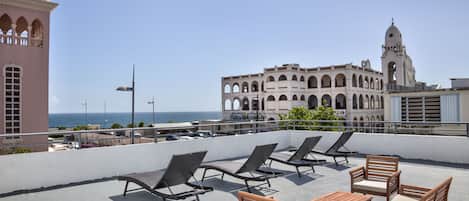 Eine Dachterrasse mit Liegestühlen und einem Tisch, mit Blick auf eine Küstenlandschaft, im Hintergrund ein historisches Gebäude und ein Turm.