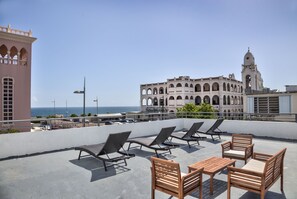 A rooftop terrace with lounge chairs and a table, overlooking a coastal area with a historic building and a tower in the background. - Casa Pelayo (San Juan)