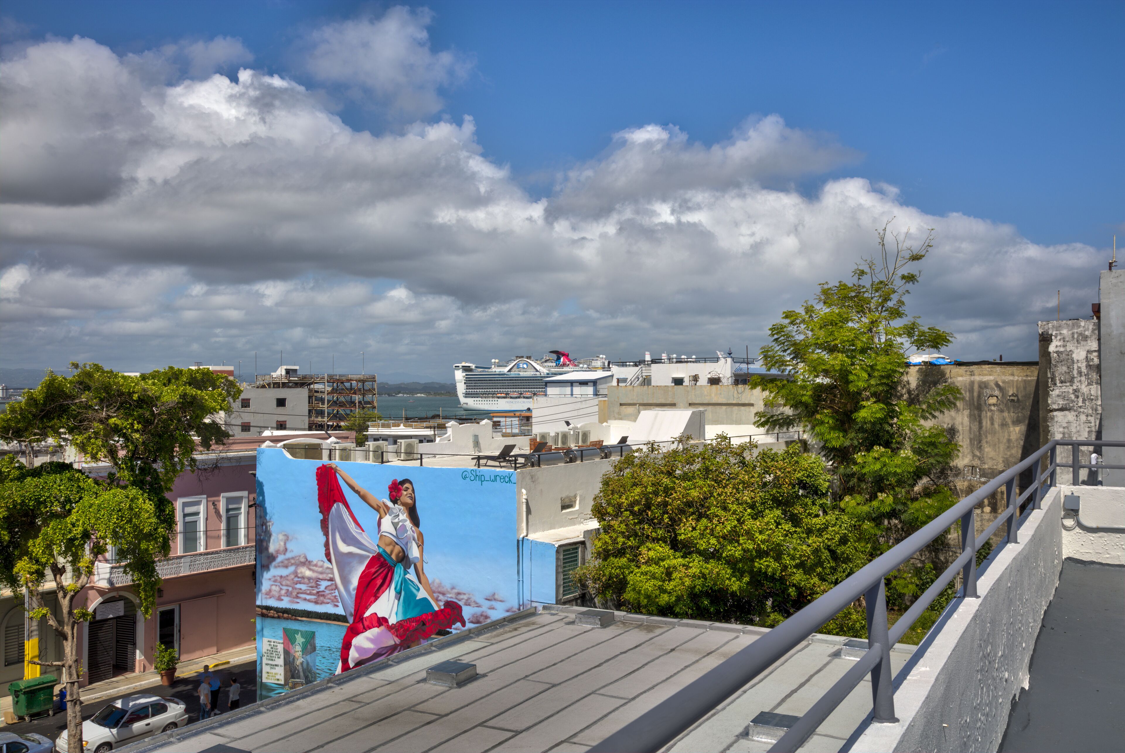 Eine Stadtansicht mit einem großen Wandgemälde an einem Gebäude, einem Kreuzfahrtschiff im Hintergrund und einem klaren, blauen Himmel.
