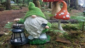 Una estatua de gnomo de jardín con un farol, en un área boscosa.