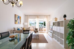 A spacious living area with a glass dining table, a sofa, a bookshelf, and a potted plant.