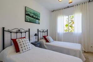 A room with two beds, a nightstand, a lamp, a painting, and a window with curtains.
